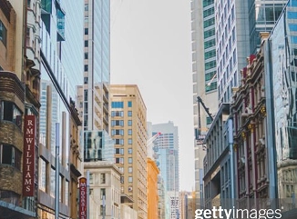 A city street scene with modern high-rise buildings and older architecture, including a visible R.M. Williams store sign on the left. The sky is clear and the image is credited to Getty Images.