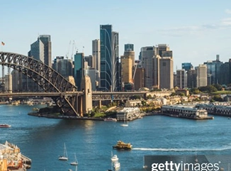 Sydney Harbour with the iconic Sydney Harbour Bridge on the left, modern city skyscrapers in the background, and boats moving across the blue water under a clear sky.