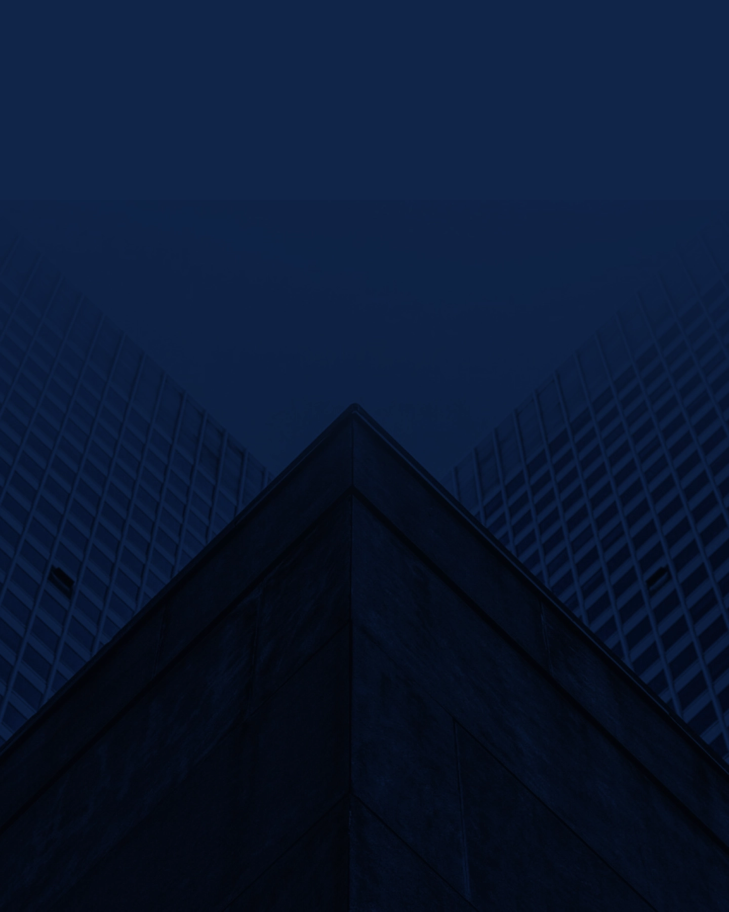 A dark blue-tinted image showing the corner of a modern building with glass and concrete facades, viewed from below, with two glass walls extending diagonally upward into the sky.